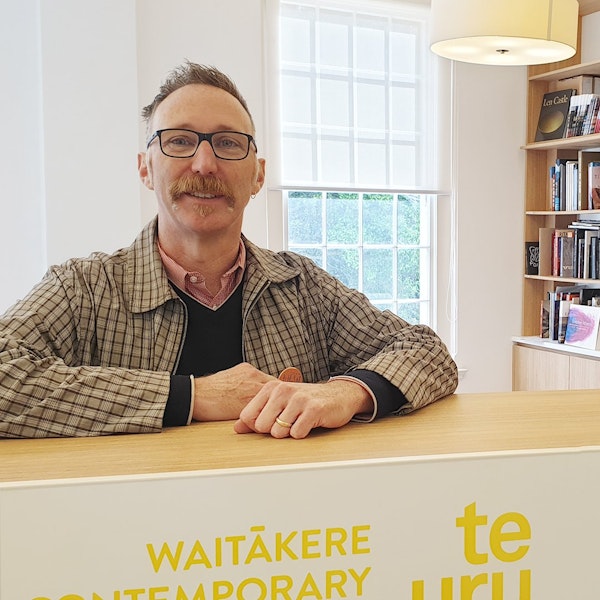 A person wearing glasses, standing behind a counter that says Te Uru Waitākere Contemporary Gallery